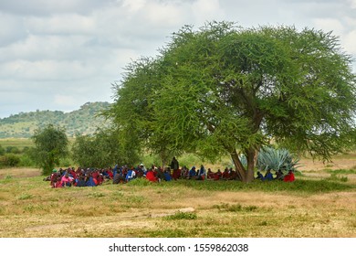 MAKUYUNI, TANZANIA -JUNE 21, 2019: African People Of The Masai Tribe In Modern, Traditional, Colorful Clothes Are Sitting Under A Tree Listening To An Elder. Makuuni Village Tribal Assembly, Tanzania