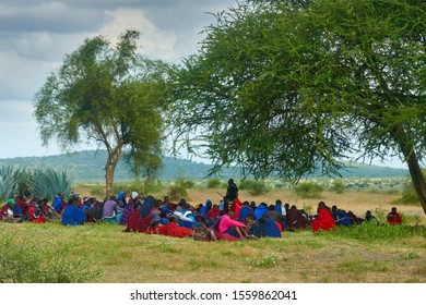 MAKUYUNI, TANZANIA - JUNE 21, 2019: African People Of The Masai Tribe In Modern, Traditional, Colorful Clothes Are Sitting Under A Tree Listening To An Elder. Makuuni Village Tribal Assembly, Tanzania