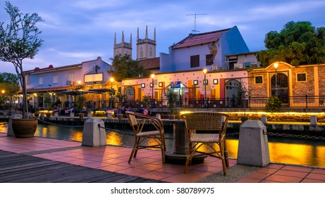 Malacca river at the evening time. Table and two chairs of street cafe on the foreground. Old church of St. Francis Xavier on the background. Light of lanterns reflecting in the river
