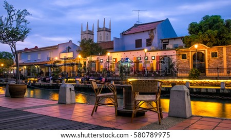 Malacca river at the evening time. Table and two chairs of street cafe on the foreground. Old church of St. Francis Xavier on the background. Light of lanterns reflecting in the river