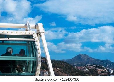 
Malaga, Spain, February 2019. People Visiting The Spanish City Of Malaga From The Cab Of The Ferris Wheel. Travel Older Couples. Beautiful Landscape: City, Mountains, Houses Against The Blue Sky.