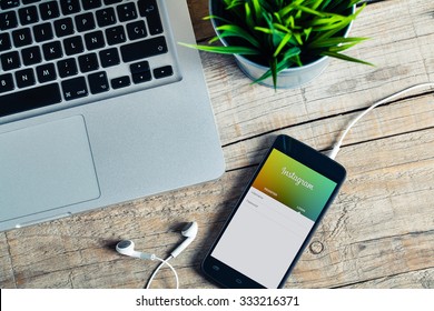 MALAGA, SPAIN - OCTOBER 29, 2015: Instagram App In A Mobile Phone Screen. Workplace With A Laptop, An Earphones And A Plant, Over A Wood Table.