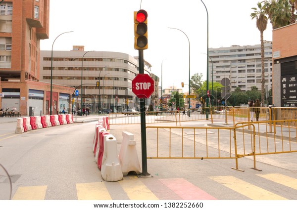 Malaga/Spain -\
04-24-2019 : Red traffic light and stop sign underneath. Yellow\
barriers, plastic red and white blocks to warn vehicles on the\
street. Buildings bordering the\
street.