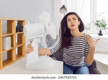 Malaise from temperature in summer. Young woman at home sits in front of stream of cold air cooling herself with electric fan. Caucasian woman feeling unwell due to high temperature during hot weather - Powered by Shutterstock