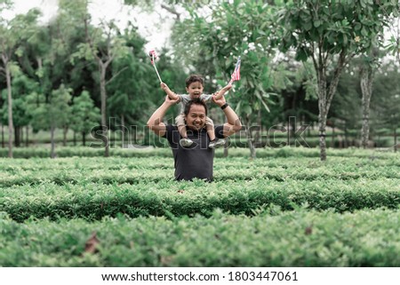 Malay boy holding Malaysia flags carried by his father on the shoulder enjoying morning sunlight at the park