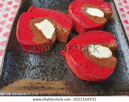 Malaysia famous cake. 3 three slices of horse shoe roll cake or Kek Tapak Kuda. Red velvet flavor with cream cheese. Soft sponge cake with filling. Black square plate. Red heart oil paper background.