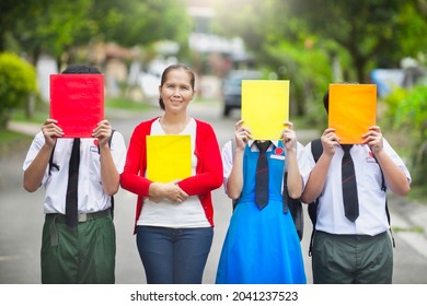 Malaysian Teacher Holding Book Standing Next To Secondary Students With Face Covered By Book.Young Asian Kids Wearing Uniform In School.Education In Asia.