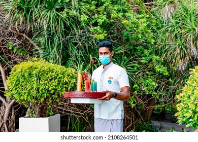 Maldives - August 2021: Maldivian Waiter, Hotel Worker In Protective Face Mask, Carries Serving Tray Of Drinks And Snacks To Swimming Pool On Luxury Tropical Island Resort