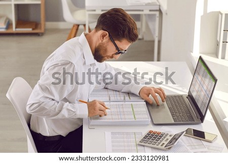 Male accountant or bookkeeper doing paperwork in the office. Young man in a white shirt and glasses sitting at his desk, using a modern laptop computer, and working with business spreadsheets