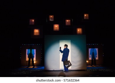 Male Actor In A Blue Suit Plays A Role In The Background Of A Theater Stage With Scenery For The Play.