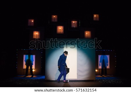 Male actor in a blue suit plays a role in the background of a theater stage with scenery for the play.