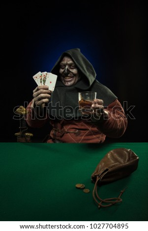 A male actor in the costume of a medieval inquisitor playing poker at a card table with a green cloth

