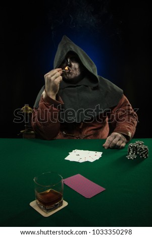 A male actor in the costume of a medieval inquisitor playing poker at a card table with a green cloth
