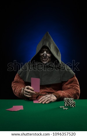A male actor in a medieval costume in a hood on his head plays poker at a card table with a green cloth