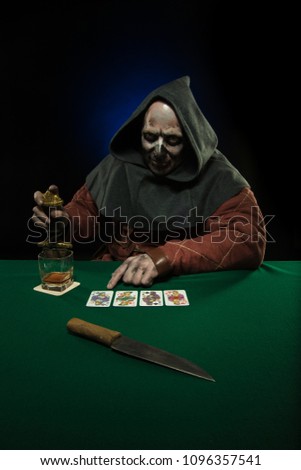 A male actor in a medieval costume in a hood on his head plays poker at a card table with a green cloth