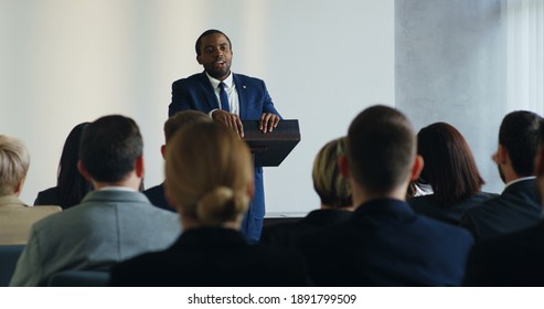 Male African American business speaker standing at the lecturn at the conference and interacting with people in the hall during speech while someone from the audience rising hand and asking the