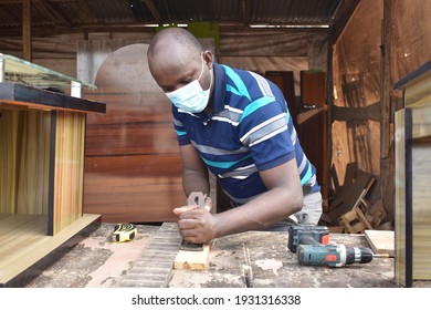 A Male African Carpenter With Nose Mask Using A Wood Plane To Smoothen A Piece Of Wood In A Carpentry Workshop  