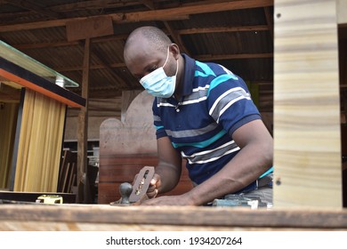 
A Male African Carpenter With Nose Mask Using A Wood Plane To Smoothen A Piece Of Wood In A Carpentry Workshop