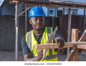 A male African construction worker with a blue safety helmet on his head, is cutting woods or planks with a short handsaw on a building site in Nigeria - Powered by Shutterstock