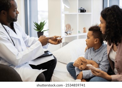 Male African doctor checks temperature of sick young African boy. Child sits on bed holding teddy bear, looking concerned. Mother, African female, sits beside, offering support. - Powered by Shutterstock