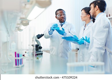 Male African-American laboratory scientist holding flask with blue liquid discussing chemical reaction with male Latin-American and female Asian colleagues. - Powered by Shutterstock