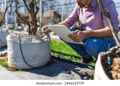 A male agriculturalist uses a tablet to analyze the development of young trees in an outdoor nursery, reflecting technology in agriculture - Powered by Shutterstock