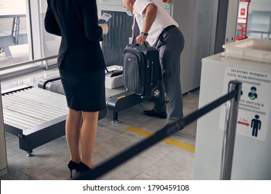 Male Airport Worker In Medical Face Mask Putting Travel Suitcase On Baggage Conveyor Belt During Security Check