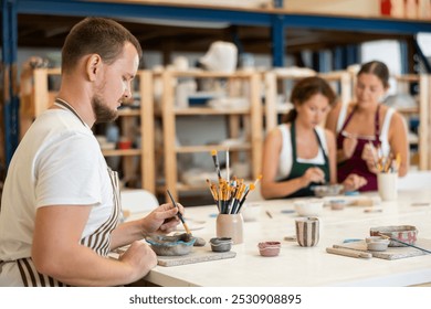 Male apprentice, is sitting near desktop and coloring clay product. In background, lovers of applied art complete design of bowl made of clay, apply pattern with brush in presence of teacher - Powered by Shutterstock