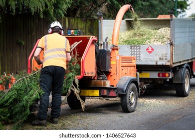 Male Arborist using a working wood chipper machine.The tree surgeon is wearing a safety helmet with a visor and ear protectors. - Shutterstock ID 1406527643