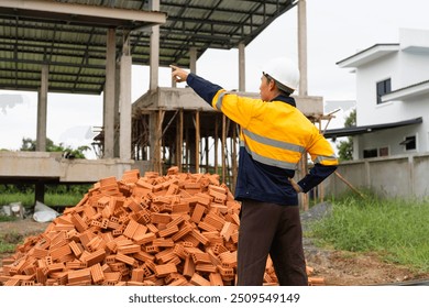 A male architect wearing a hard hat and safety vest checks his laptop while holding a clipboard with house plan papers. He reviews blueprints, construction documents,house structure details on-site. - Powered by Shutterstock