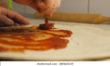 Male arm of cook applying ketchup on pizza dough using a spoon at kitchen table. Hand of chef spreading tomato sauce on pastry in metal form on a wooden surface at cuisine. Concept of preparing food