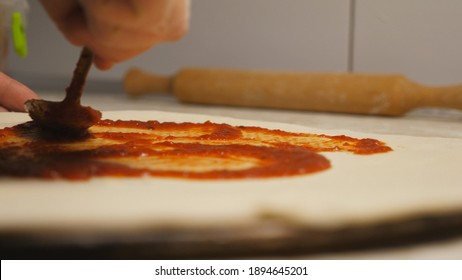 Male arm of cook applying ketchup on pizza dough using a spoon at kitchen table. Hand of chef spreading tomato sauce on pastry in metal form on a wooden surface at cuisine. Concept of preparing food