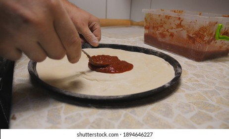 Male arm of cook spreading tomato sauce on pizza dough using a spoon at kitchen table. Hand of chef applying ketchup on pastry in metal form at wooden surface. Concept of preparing food. Close up