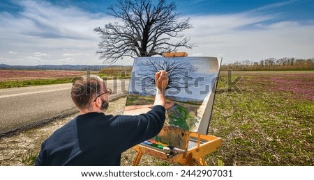 Male artist paints large lonely oak tree growing in side of rural road early in spring; fields and distant forest in background