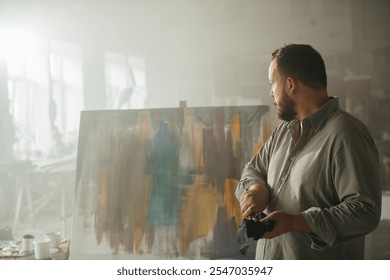 Male artist thoughtfully looking at a freshly completed painting in his studio. - Powered by Shutterstock