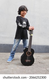  Male Asian Kid Or A Young Boy With Long Hair Pose Wearing Black T Shirt Also Jeans And Holding A Black Mini Guitar 