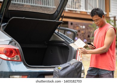 Male Asian Resident Wearing Red Shirt At Sydney South Standing Checking His Mobile Phone And Reviewing Bushfire Emergency Rescue Plan Prior Lifting Luggage Into Car Hood And Leaving The House