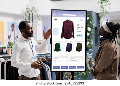 Male assistant showing clothes on interactive kiosk board to young trendy customer in clothing store at mall. Retail employee helping client to choose modern fashion items, self ordering. - Powered by Shutterstock