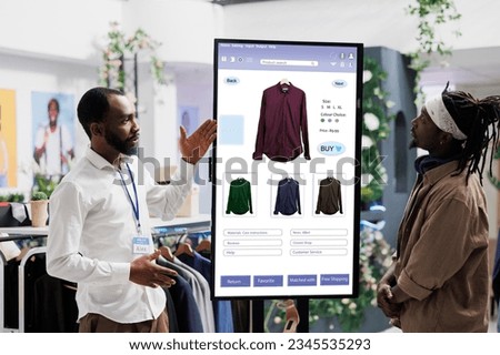 Male assistant showing clothes on interactive kiosk board to young trendy customer in clothing store at mall. Retail employee helping client to choose modern fashion items, self ordering.