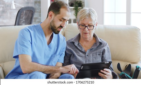 Male assistant teaching senior retired elderly old woman how to use her tablet computer in nursing home for her therapy