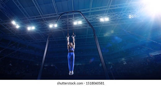 Male Athlete Doing A Complicated Exciting Trick On Gymnastics Rings In A Professional Gym. Man Perform Stunt In Bright Sports Clothes