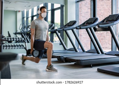 Male Athlete Exercising With Hand Weights In Lunge Position While Wearing Protective Face Mask In Health Club. 