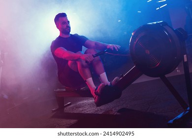 A male athlete pulls the oars on a rowing machine, his form lit by moody blue and red lights, shrouded in light fog - Powered by Shutterstock