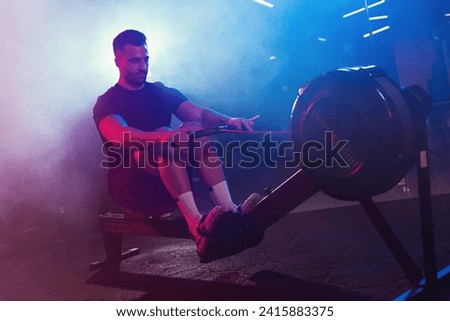 A male athlete pulls the oars on a rowing machine, his form lit by moody blue and red lights, shrouded in light fog
