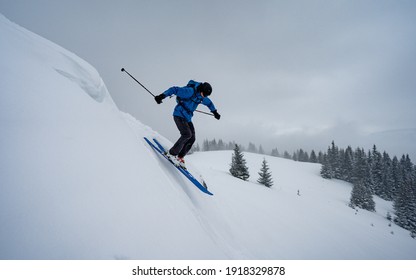 A Male Athlete Skier In Blue Ski Jacket Makes A Jump With Flying Snow Powder.  Winter Extreme Sports Concept. Snow Falling From Heavy Grey Clouds, Windy Cold Weather.