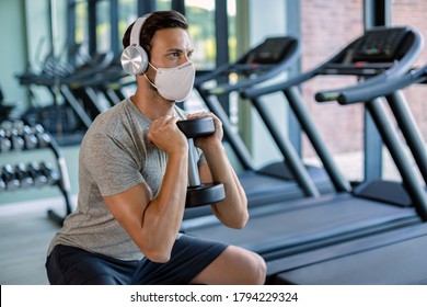 Male Athlete Using Hand Weight While Doing Squats And Wearing Protective Face Mask In Health Club. 