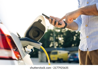 Male attaching in power cord to electric car using app on smartphone. Man plugging electric car from charging station. Businessman charging electric car on charging station at sunset