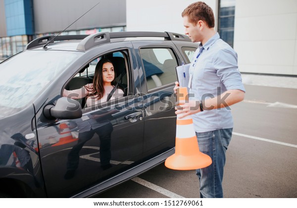 Male
auto instructor takes exam in young woman. Stand outside of car
with orange sign in hands. Look at woman in car. Female studenthold
hands on steering wheel adnd look at
instructor.