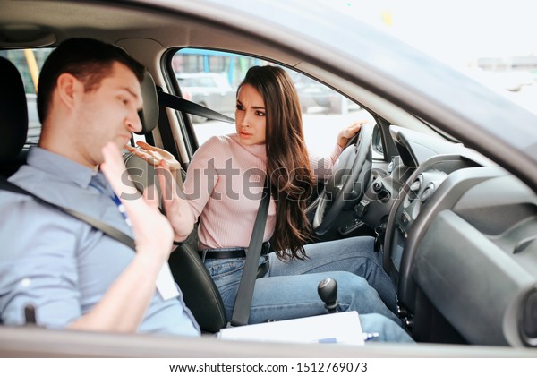 Male auto instructor takes exam in\
young woman. Female student look at teacher with confusion and\
misunderstanding. Guy show hands and look down.\
Daylight.