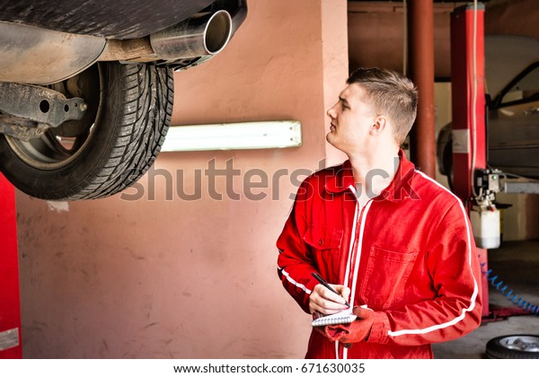Male auto mechanic standing making notes\
underneath a lifted car near working equipment for repair and\
diagnostic in auto repair\
service